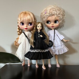 May include: Three fashion dolls are posed together. The dolls have large eyes and are wearing dresses. The doll on the left has blonde hair and a white dress. The center doll has long brown hair and a black dress. The doll on the right has curly blonde hair and a white dress.