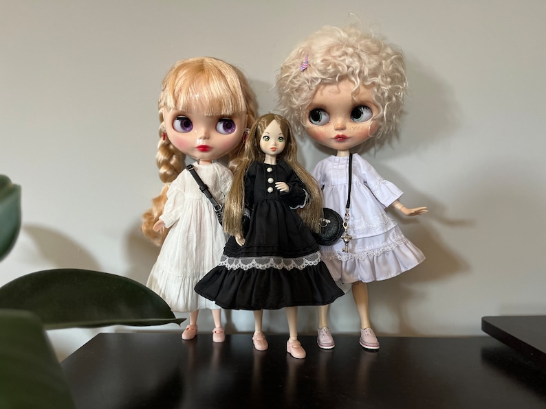 May include: Three fashion dolls are posed together. The dolls have large eyes and are wearing dresses. The doll on the left has blonde hair and a white dress. The center doll has long brown hair and a black dress. The doll on the right has curly blonde hair and a white dress.