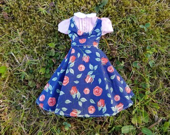 May include: A doll dress featuring a navy blue pinafore with a rose pattern and a light pink blouse. The dress has a white collar and is set against a backdrop of green grass.