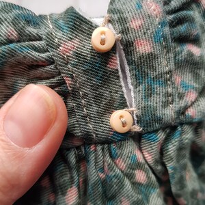 May include: Close-up of a green and blue floral fabric with two white buttons. The buttons are sewn to the fabric and are connected by a white thread.