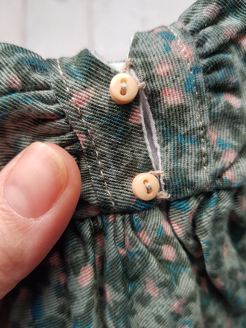 May include: Close-up of a green and blue floral fabric with two white buttons. The buttons are sewn to the fabric and are connected by a white thread.