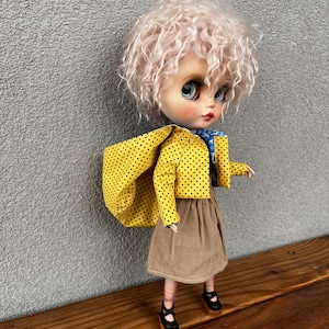 Può includere: Bambola con parrucca riccia rosa chiaro, indossa una giacca gialla a pois, una gonna di velluto a coste marrone e scarpe Mary Jane nere. La bambola ha grandi occhi blu e si trova su una superficie di legno.