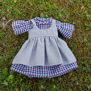 May include: A doll's dress set, featuring a collared shirt with a red, white, and blue plaid pattern, topped with a gray pinafore dress. The outfit is displayed on a bed of green grass.