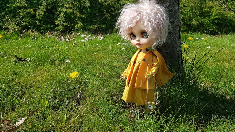 May include: A doll with pink curly hair wearing a yellow dress and a green jacket stands in a grassy field. The doll is holding a small, round, yellow object in its hand.