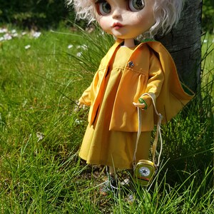 May include: A doll with white curly hair wearing a yellow dress and jacket, standing in a grassy field.