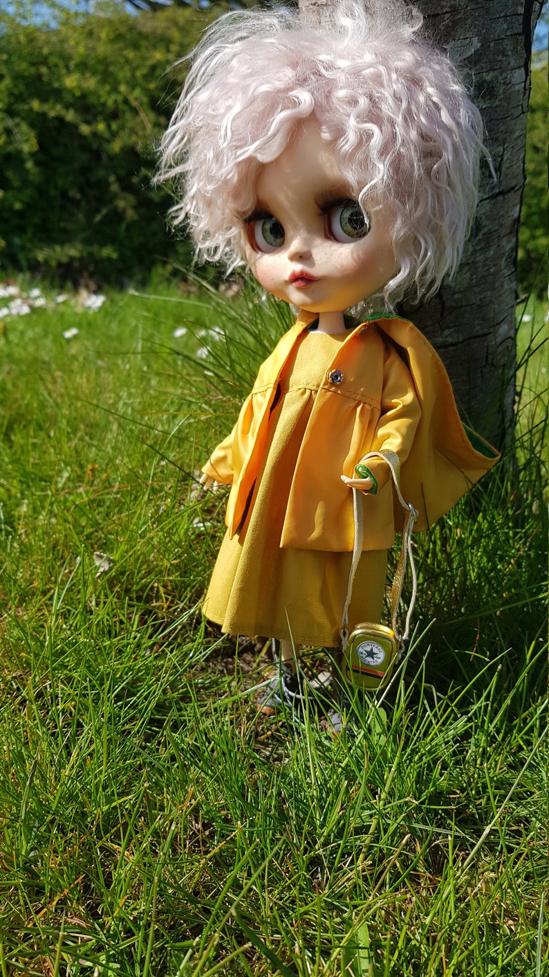 May include: A doll with white curly hair wearing a yellow dress and jacket, standing in a grassy field.