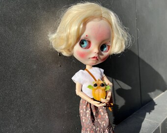 Puede incluir: Una muñeca con cabello rubio, ojos azules y mejillas rosadas, que lleva una camiseta blanca y una falda estampada. La muñeca sostiene un pequeño bolso naranja con forma de búho con una correa marrón. El fondo es una pared gris oscuro.