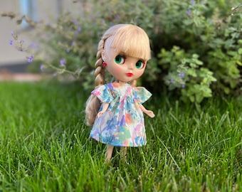 Puede incluir: Una muñeca con el pelo rubio trenzado lleva un vestido floral y está de pie en un campo de hierba.