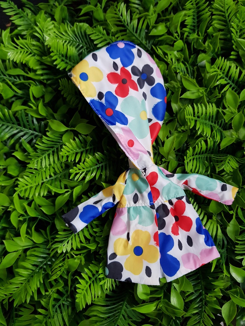 May include: A white raincoat with a hood, featuring a colorful floral pattern with blue, red, yellow, and green flowers. The raincoat is made of a lightweight fabric and has a relaxed fit.