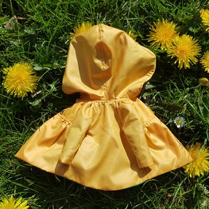 May include: A yellow hooded raincoat with a green lining, lying on a bed of green grass with yellow dandelions.