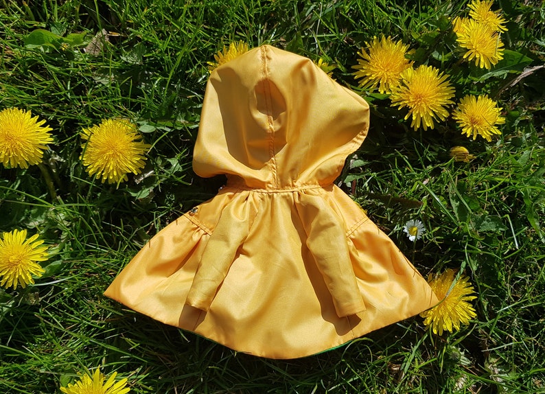 May include: A yellow hooded raincoat with a green lining, lying on a bed of green grass with yellow dandelions.