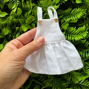 May include: White linen doll's dress with a ruffled hem and two gold buttons. The dress has a simple, classic design.
