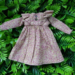 May include: A beige doll dress with a pink floral pattern and ruffled sleeves. The dress has a gathered waist and a button closure at the neckline.