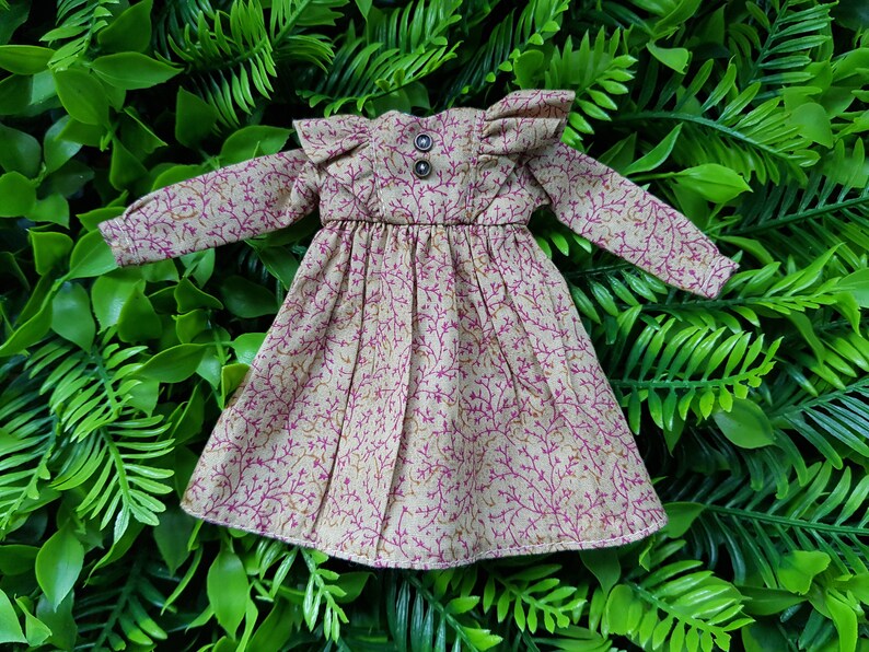 May include: A beige doll dress with a pink floral pattern and ruffled sleeves. The dress has a gathered waist and a button closure at the neckline.