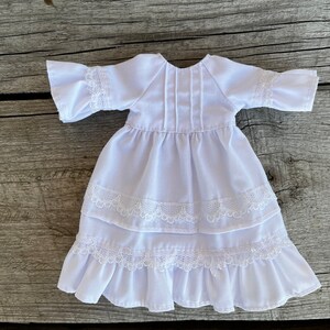 May include: A white doll dress with ruffled sleeves and a tiered skirt. The dress features delicate lace trim on the sleeves and skirt layers. The dress is made of a lightweight fabric.
