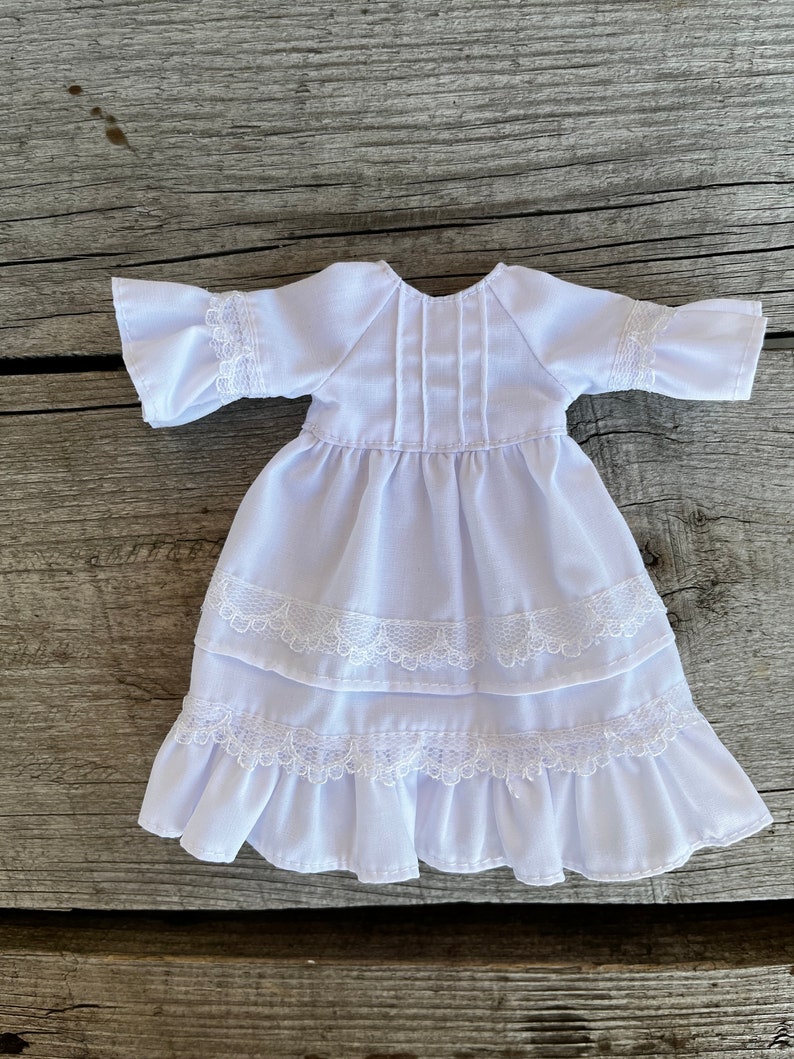 May include: A white doll dress with ruffled sleeves and a tiered skirt. The dress features delicate lace trim on the sleeves and skirt layers. The dress is made of a lightweight fabric.