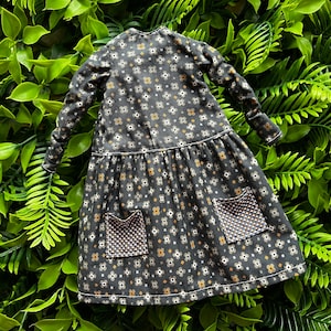Puede incluir: Un vestido en miniatura con mangas largas y falda acampanada. El vestido es negro con un estampado floral en blanco, marrón y dorado. Tiene dos bolsillos delanteros con un patrón geométrico.