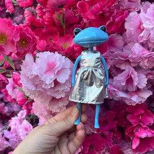 Könnte beinhalten: Eine blaue Froschfigur mit einem silbernen Kleid, vor einem Hintergrund aus rosa und magenta Blüten. Die Figur hat einen runden Kopf, schwarze Augen und blaue Gliedmaßen. Das Kleid ist metallisch und glänzend.