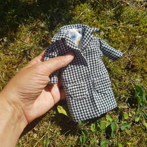 May include: A miniature black and white houndstooth coat with a collared neckline and two front pockets. The coat's interior lining features a floral pattern. The coat is held against a backdrop of green grass and foliage.