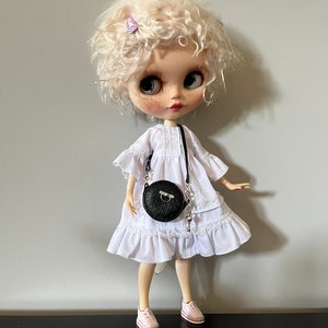 May include: A doll with curly blonde hair, wearing a white dress with lace trim, a black round purse, and pink sneakers. The doll has large eyes and a small purple hair clip. The doll is standing on a black surface against a gray background.