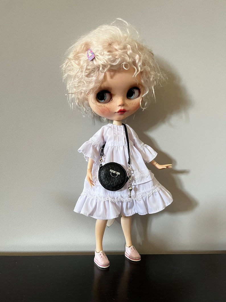 May include: A doll with curly blonde hair, wearing a white dress with lace trim, a black round purse, and pink sneakers. The doll has large eyes and a small purple hair clip. The doll is standing on a black surface against a gray background.