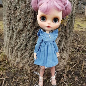 May include: A doll with pink hair in pigtails wearing a blue denim dress and brown boots. The doll is standing in front of a tree trunk.