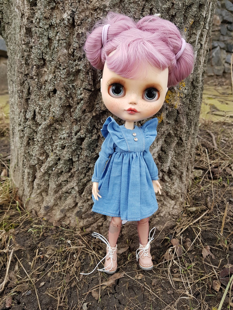 May include: A doll with pink hair in pigtails wearing a blue denim dress and brown boots. The doll is standing in front of a tree trunk.