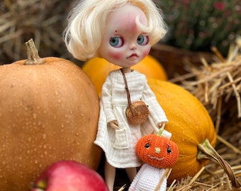 May include: A blonde doll wearing a white dress and holding a crocheted pumpkin toy. The doll is surrounded by pumpkins and hay.