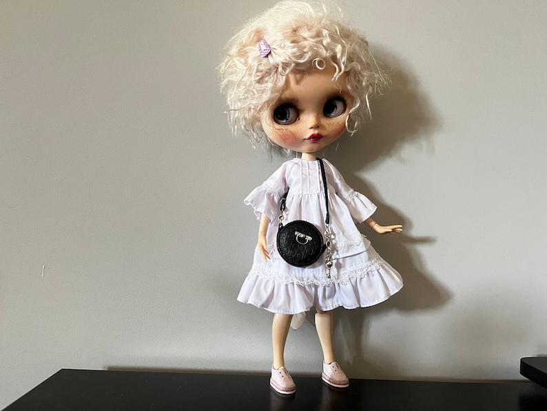 May include: A doll with curly blonde hair, wearing a white dress and pink shoes. The doll has a black round purse with a silver chain. The doll has a fair complexion with rosy cheeks and lips. A small purple hair clip is in the doll's hair.