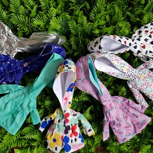 May include: A collection of seven doll-sized hooded raincoats in various colors and patterns, including silver, blue with stars, turquoise, pink with watermelon print, white with floral print, and white with red hearts.