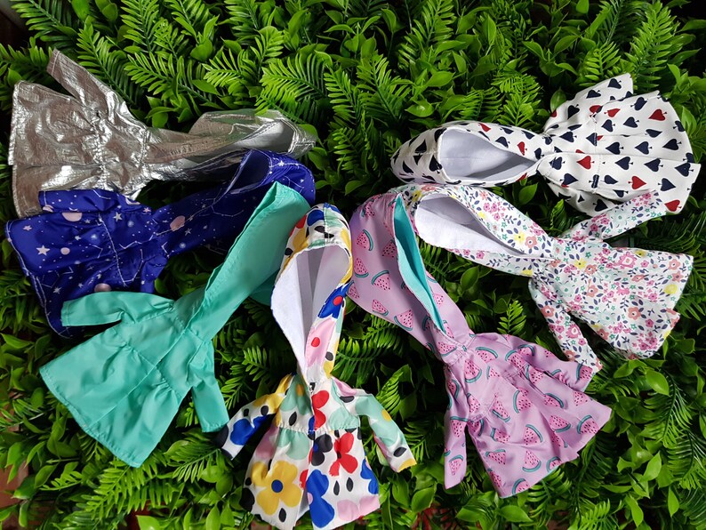 May include: A collection of seven doll-sized hooded raincoats in various colors and patterns, including silver, blue with stars, turquoise, pink with watermelon print, white with floral print, and white with red hearts.