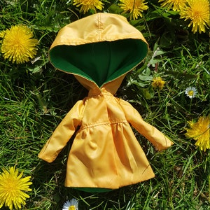 May include: A yellow raincoat with a green hood, lying on a bed of green grass with yellow dandelions.