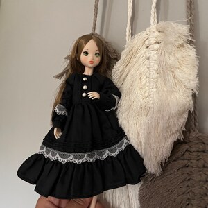May include: A doll with long brown hair and green eyes wearing a black dress with white lace trim and pearl buttons. The doll is posed in front of a macrame wall hanging with cream and brown accents.