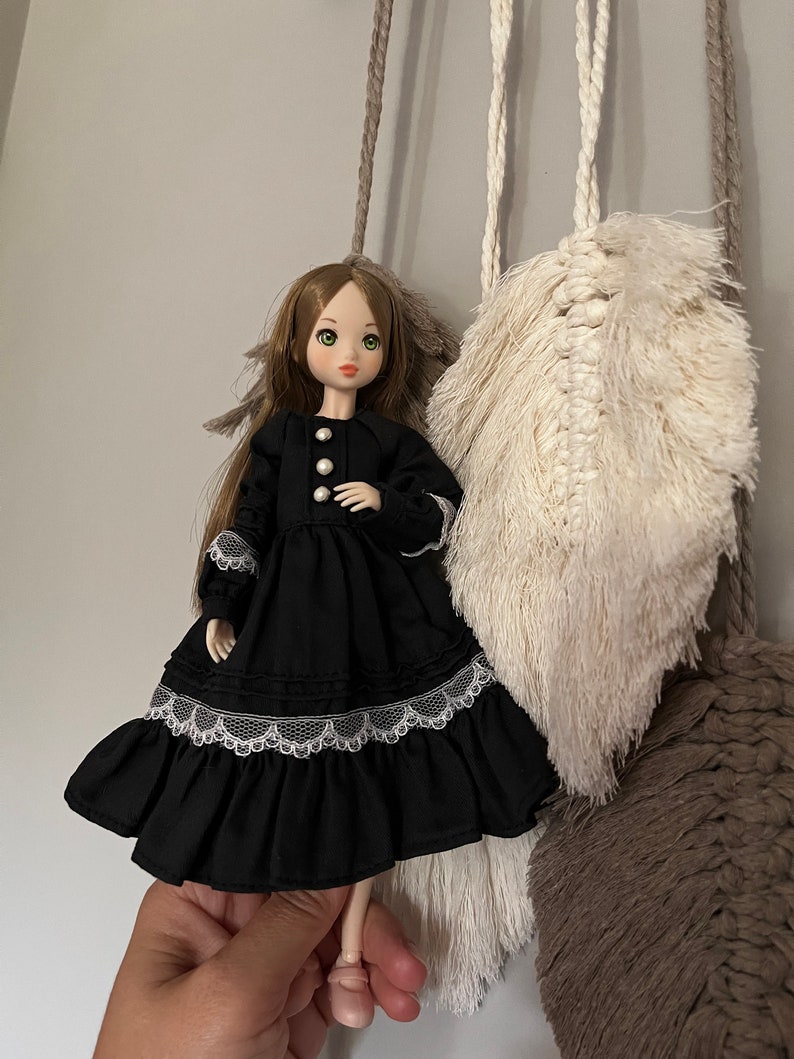 May include: A doll with long brown hair and green eyes wearing a black dress with white lace trim and pearl buttons. The doll is posed in front of a macrame wall hanging with cream and brown accents.