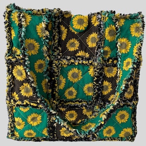 May include: A quilted tote bag with a sunflower pattern. The bag features a patchwork design with green and brown squares, each adorned with yellow sunflowers. The edges are fringed, and a sunflower button secures the top.