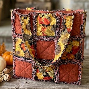 May include: A quilted tote bag constructed from square fabric patches featuring a mix of solid burnt orange and patterned panels with sunflowers and autumn leaves. The bag has frayed, exposed seams between the squares and two fabric handles.