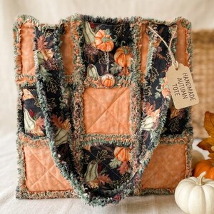 May include: A quilted tote bag featuring a patchwork design of solid orange squares and black fabric panels printed with pumpkins and autumn leaves. The bag has frayed seams and a beige tag attached with string that reads HANDMADE AUTUMN TOTE.