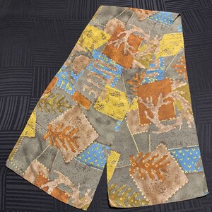 Puede incluir: Una bufanda de tela con estampado gris y marrón con un diseño de patchwork. La bufanda presenta varias formas y colores, incluyendo amarillo, azul y marrón. La tela tiene un aspecto texturizado.