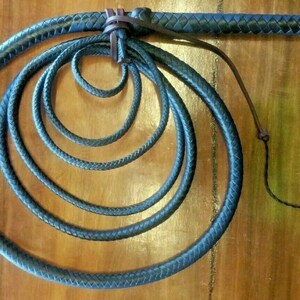 Puede incluir: L&aacute;tigo de cuero azul oscuro con un mango trenzado y una serie de bucles conc&eacute;ntricos. El l&aacute;tigo est&aacute; hecho de un material azul oscuro y se muestra sobre un fondo de madera. El mango est&aacute; envuelto en cuero marr&oacute;n m&aacute;s oscuro.
