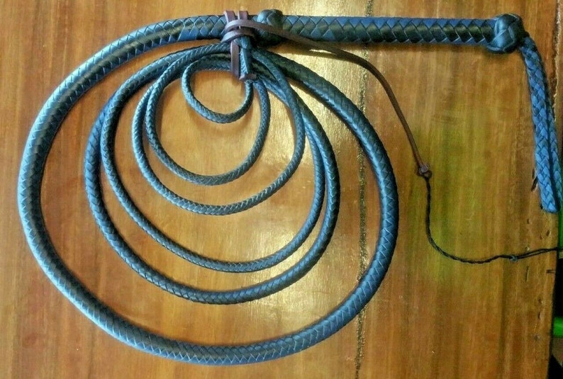 Puede incluir: L&aacute;tigo de cuero azul oscuro con un mango trenzado y una serie de bucles conc&eacute;ntricos. El l&aacute;tigo est&aacute; hecho de un material azul oscuro y se muestra sobre un fondo de madera. El mango est&aacute; envuelto en cuero marr&oacute;n m&aacute;s oscuro.