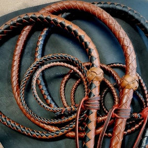 May include: Close-up of three braided leather whips with brown and black leather. The whips are coiled together and have a decorative knot at the end.