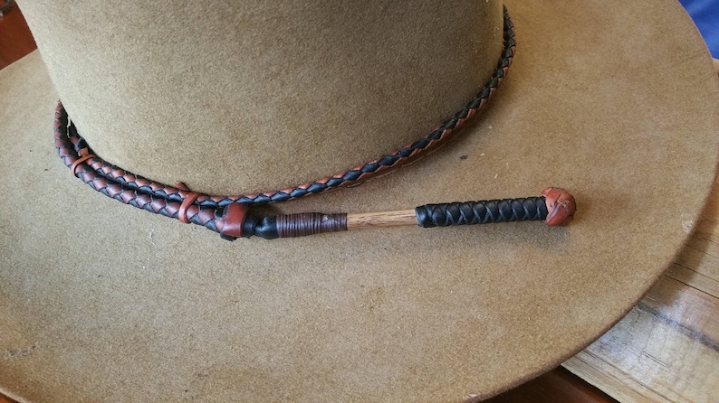 Op de afbeelding: Een bruine leren hoed met een zwart en bruin gevlochten leren hoedband. De hoedband heeft een houten stok met een zwart en bruin gevlochten leren omhulsel en een bruine leren punt.