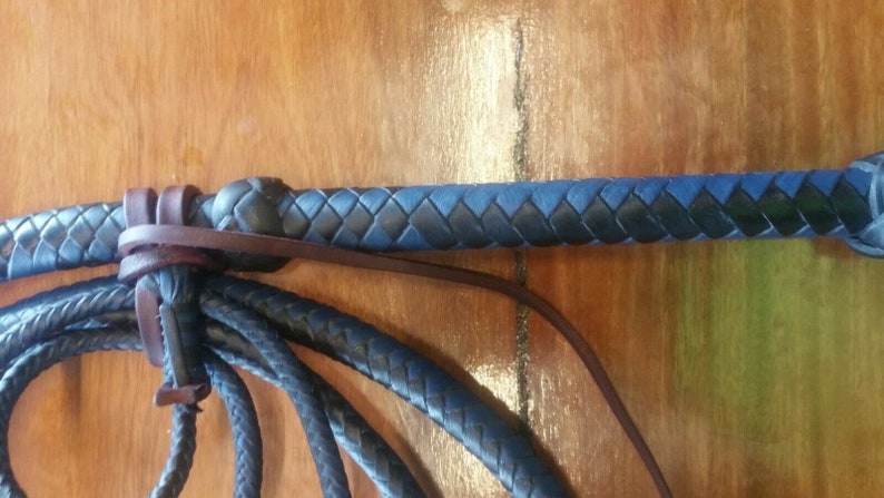 Puede incluir: Un l&aacute;tigo de cuero negro y azul con un mango trenzado y un cuerpo trenzado. El l&aacute;tigo est&aacute; hecho de cuero y tiene una correa de cuero marr&oacute;n. El l&aacute;tigo est&aacute; sobre una superficie de madera.