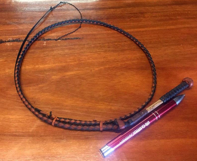 Op de afbeelding: Een bruine en zwarte gevlochten leren zweep met een houten handvat en een rode pen met de tekst "Just Saying" erop.