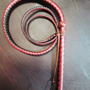 May include: A red and black leather bullwhip with a braided handle. The whip is coiled and ready to be used.