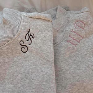 May include: Two light gray heathered sweatshirts featuring ribbed mock neck collars. The left sweatshirt displays the embroidered black initials SK, while the right sweatshirt features the embroidered pink initials HD on the upper chest area.