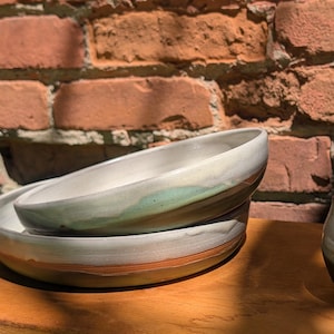 May include: Two stacked ceramic bowls with a smooth white interior and a light green glaze accent on the rim, paired with a small, rounded ceramic cup featuring a speckled white upper section and a solid dark brown base, all with a hand-thrown finish.
