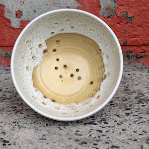 May include: A handmade ceramic bowl with a white exterior and a tan interior. The bowl has numerous small holes around the rim and in the base. The bowl is sitting on a concrete surface with a red and peeling paint background.
