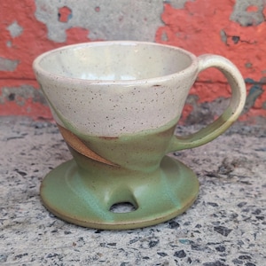May include: Handmade ceramic pour-over coffee maker. The top is speckled off-white, transitioning to a light green base. Features a handle and a circular base with a hole. The design includes a brown accent. Perfect for brewing coffee.