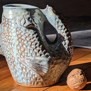 May include: A ceramic jug with a fish design, featuring a light blue glaze and brown accents. The jug has a handle and a textured surface. A walnut sits beside the jug on a wooden surface.